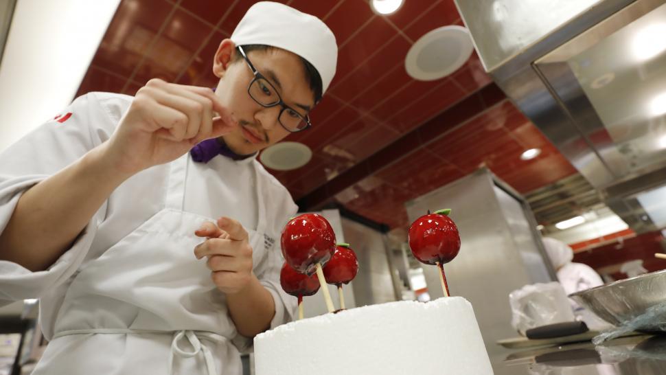 A person wearing a chef’s uniform sprinkles something over a dessert that looks like candied apples.