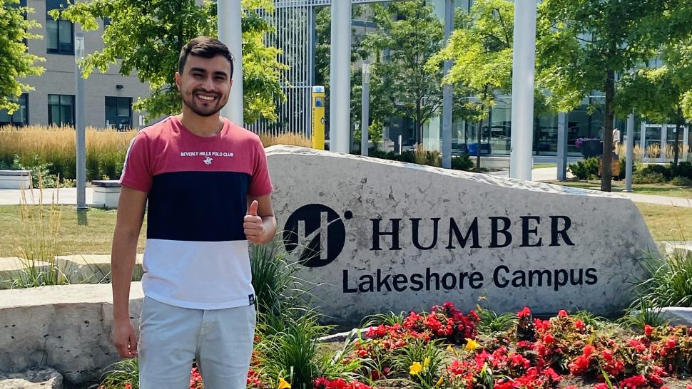 A smiling person stands in front of a sign the reads Humber Lakeshore Campus that sits in a flower bed.