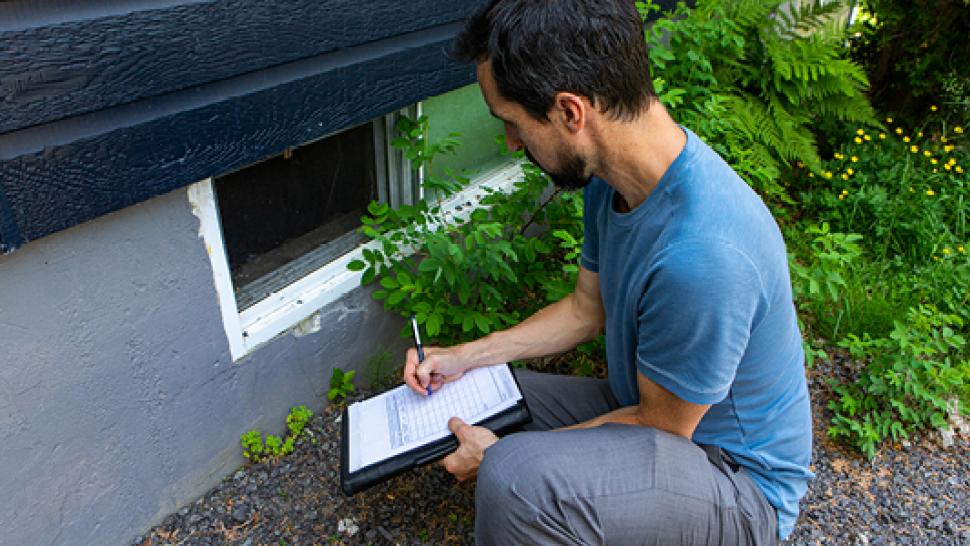 A person holding a clipboard is looking at a window on a home.