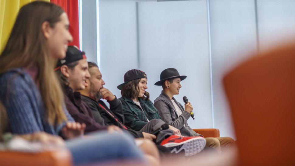 A group of five smiling people sit in a row on a stage. One is speaking into a microphone.