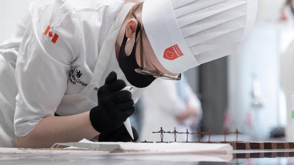 Emma Kilgannon wears her chef’s uniform while using a pastry bag to create small chocolate figures for a dessert.