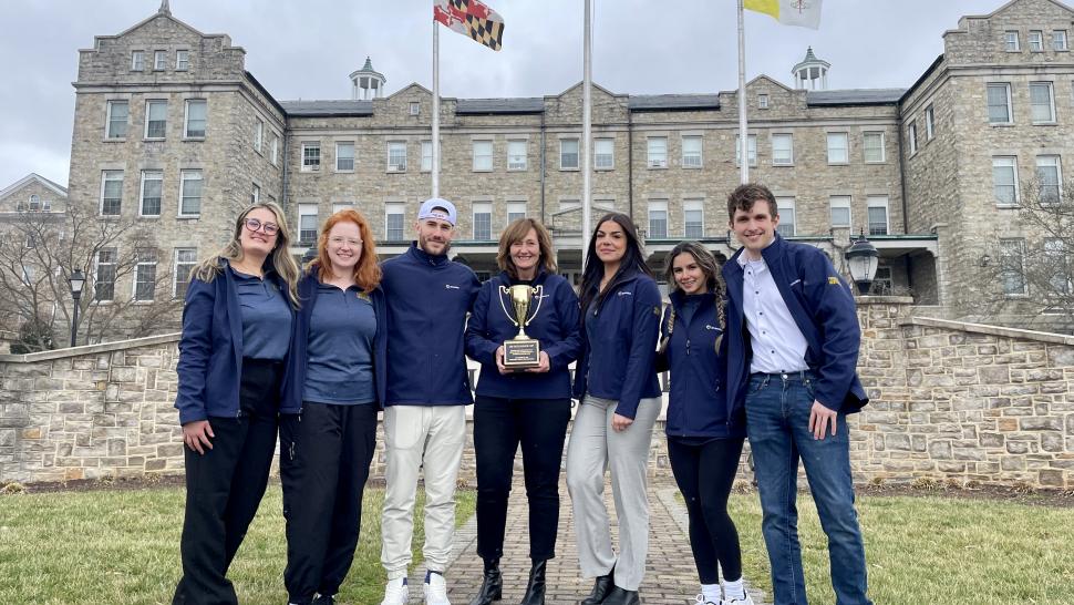 Humber College students finished first and second in the Robert Fram CSI Challenge. Seven people stand arm-in-arm in front of a building. The person in the middle is holding a gold trophy.