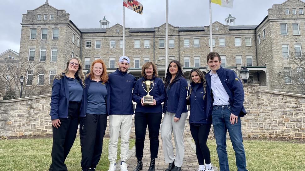 Seven people stand arm-in-arm in front of a building. The person in the middle is holding a gold trophy.