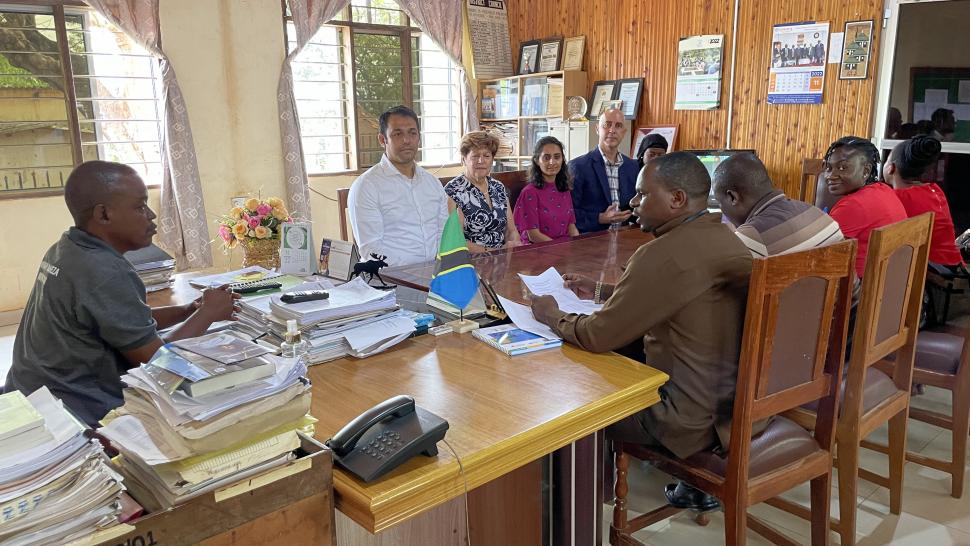 Humber College is helping strengthen alternative pathways to education and employment for women and adolescent girls in Tanzania. A group of people sit around a table filled with papers and books while one person addresses them.
