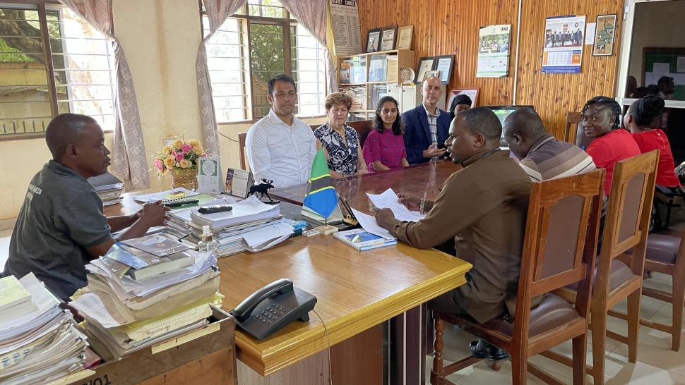 A group of people sit around a table filled with papers and books while one person addresses them.