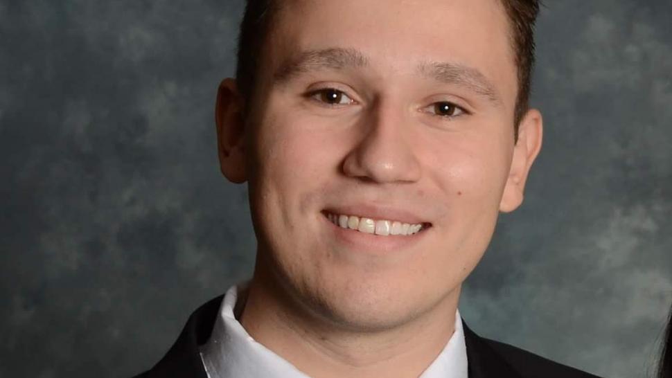 Paul-Michael Broz smiles in front of a grey background, wearing a black tie and jacket and white button down