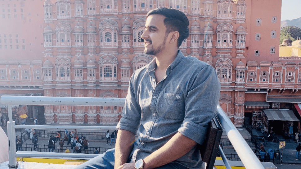 Kanishk Bhuria is seated, looking over his right shoulder with a smile, an old building with a beautiful facade in the backgrogr