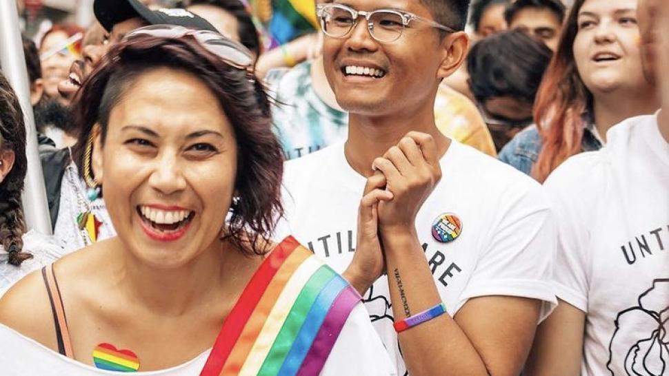 Kathryn Mettler (left) and Michael Butac smile widely, surrounded by bright colours and people at a Pride parade