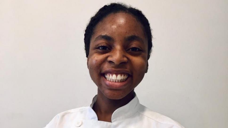 Angelique Ricketts is dressed in a chef's coat and is holding a jar of preserves