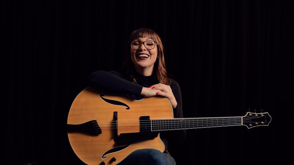 A smiling woman holding a guitar and sits in front of a black backdrop.