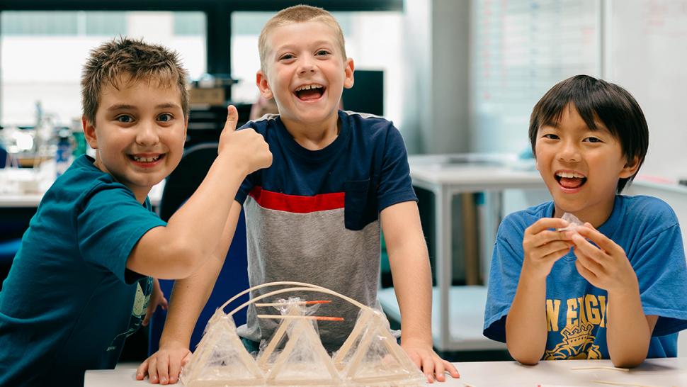 The Barrett STEAM Academy empowers students to think critically, solve real-world challenges, and build future-ready skills through project-based learning. Three smiling youth are building a miniature bridge using sticks.