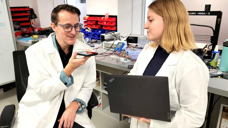 Two people are wearing lab coats. One is holding a smartphone and the other a laptop.