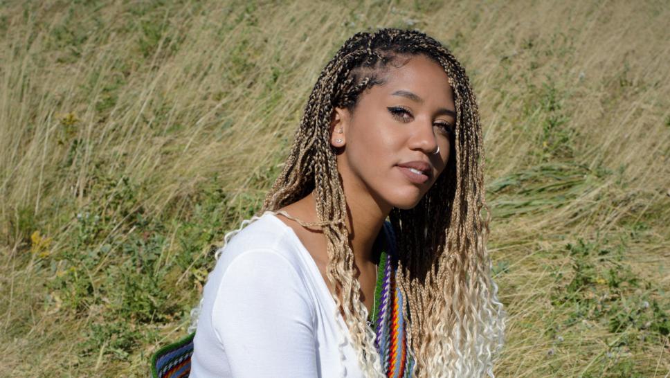 Larissa Crawford peers into the camera from a crouched but powerful position. She is wearing a shirt, jeans and colourful sash