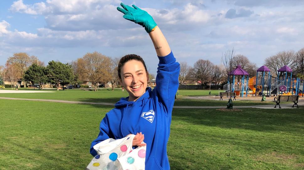 Marianna Butelli is holding a polka dot trash bag and wearing gloves with one arm raised in celebration in a green field 