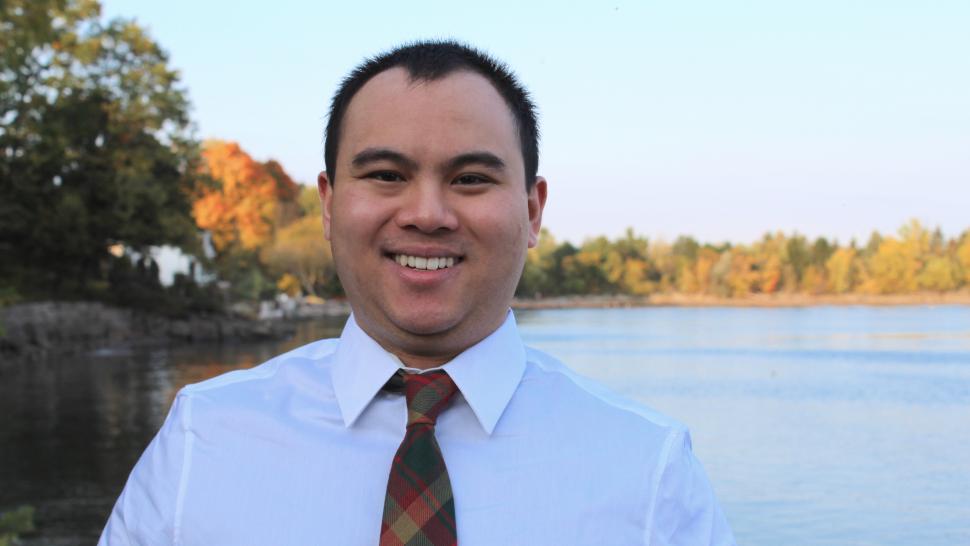 PR student Mathias Ho smiles, wearing a white shirt and red tie. There is a lake surrounded by colorful trees in the background