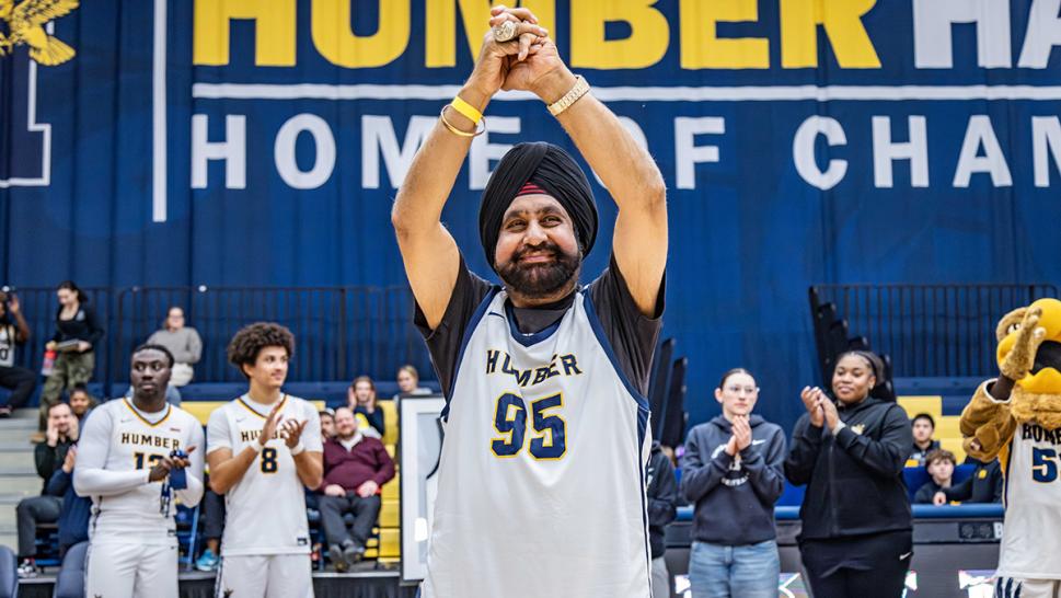 A person wearing a Humber Hawks jersey raises their hands above their head as others in the background clap.