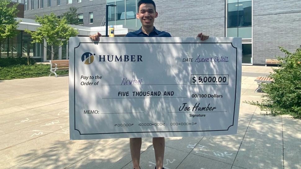 Newton Lew holds up a giant cheque for $5,000 from Humber College which hides most of his body.He's wearing a t-shirt and shorts