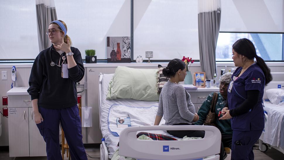 Nurses stand next to a hospital bed that a patient is sitting on.