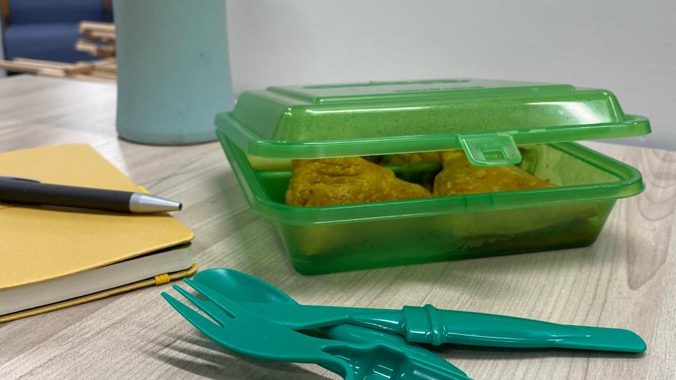 A reusable container with food inside it sits on a desk with a reusable water bottle and reusable cutlery near it.