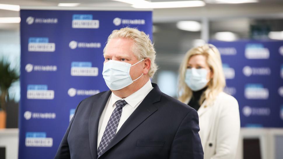 Premier Doug Ford is seen wearing a blue surgical mask and black suit at photo op. Natalia Kusendova is seen in the background.