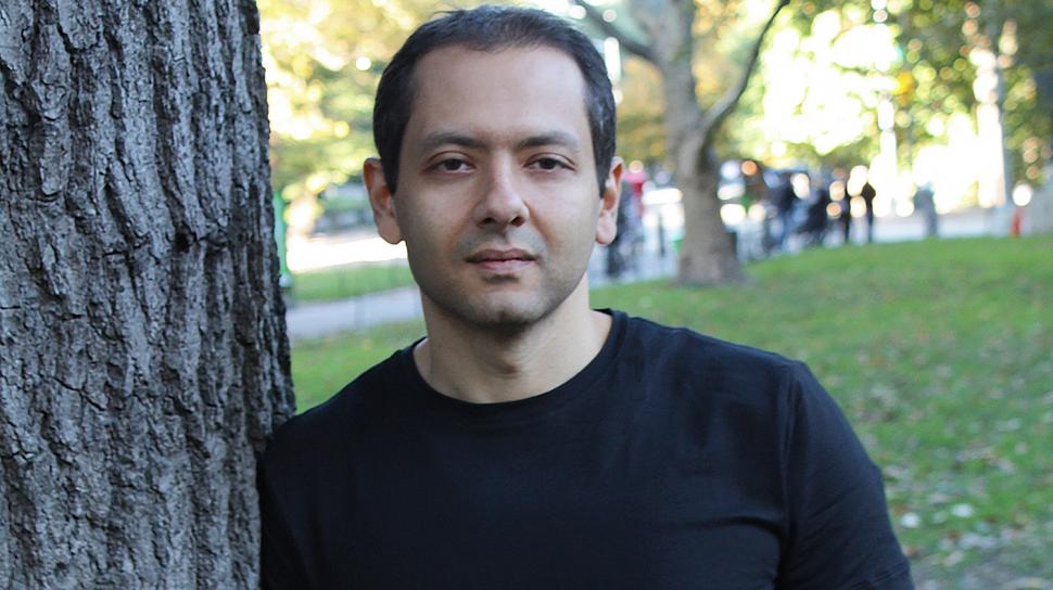 Omar El Akkad leans against a tree in a grassy park wearing a black t-shirt and jeans, hands in pockets