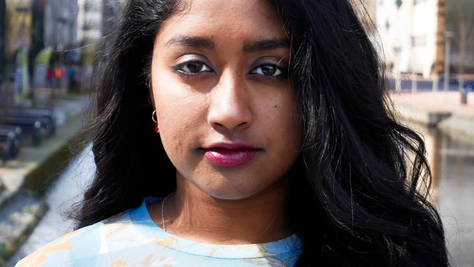 Svedha Krishnaraj smiles slightly in front of a cityscape on a sunny day. She has long dark hair and wears berry lipstick