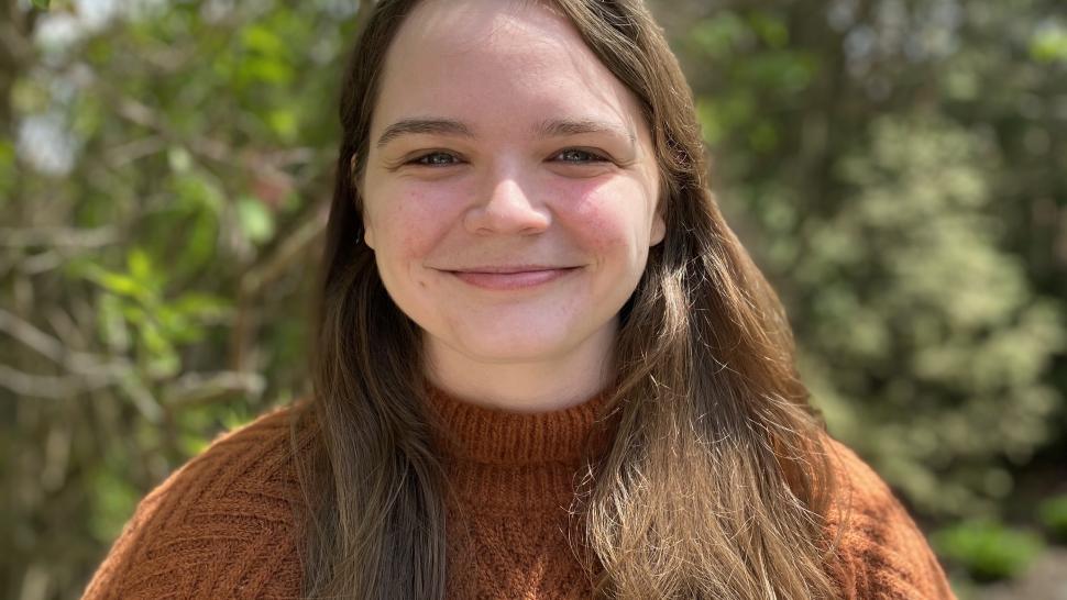 Anne Campbell smiles, close-mouthed, in front of green foliage. She is wearing a rust-coloured sweater and has long brown hair