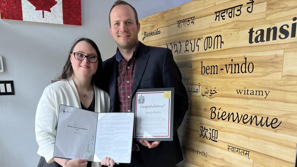 Two people pose for a photo holding documents. They stand in front of a small Canadian flag and a wooden backdrop.