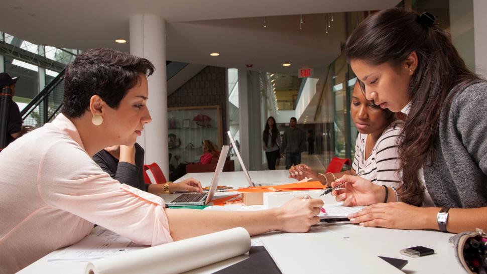 A person sitting at a table uses a pen to point to a section on a piece of paper as two other people look at the paper. 