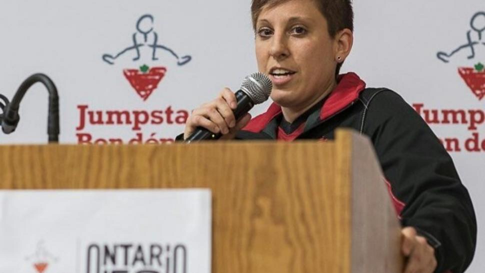 Claire Buchanan speaks at a podium wearing her national team jacket, holding a microphone.