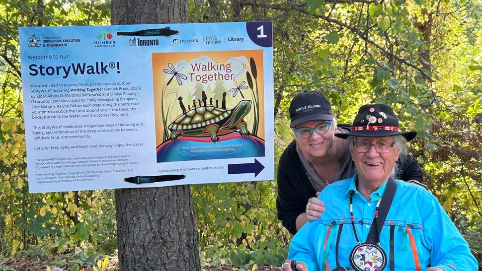 Two people are in a forest beside a sign that reads StoryWalk.