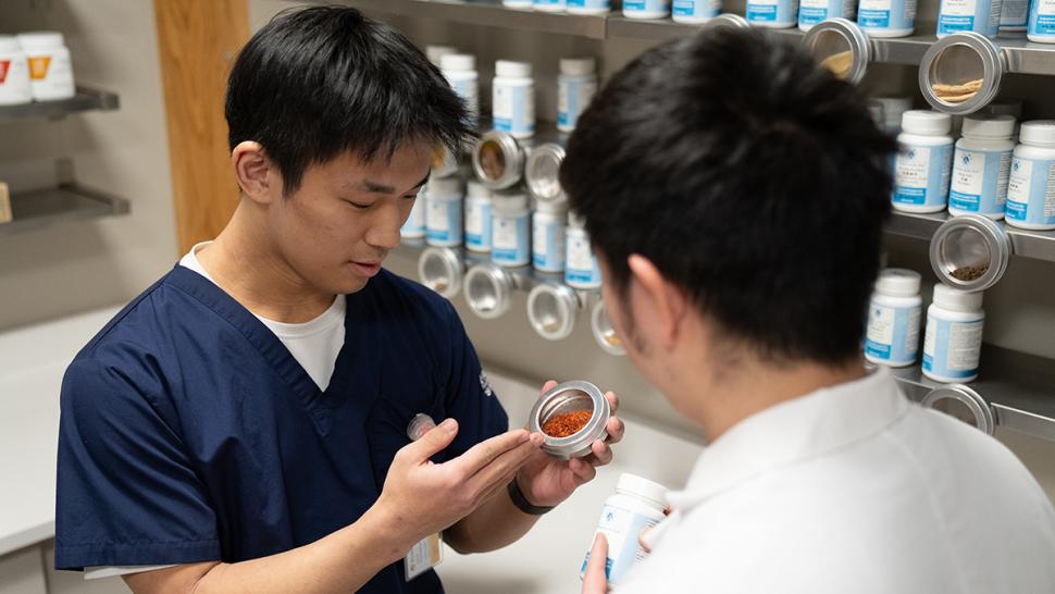 Two people are talking and one is holding a jar of what appears to be dried herbs.