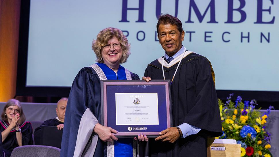 Humber Polytechnic conferred an honorary degree on Ted Nolan as part of the Spring 2025 Convocation ceremonies. Two people wearing graduation robes stand together and are holding an honorary degree.