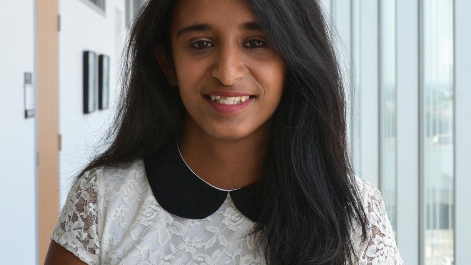 Treta Vyas stands in a hallway lined with windows in Humber College's LRC building. She is wearing a white T-shirt and smiling