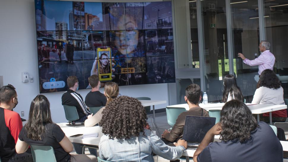 Students listen while a person looks at a screen with images on it and talks into a microphone. 