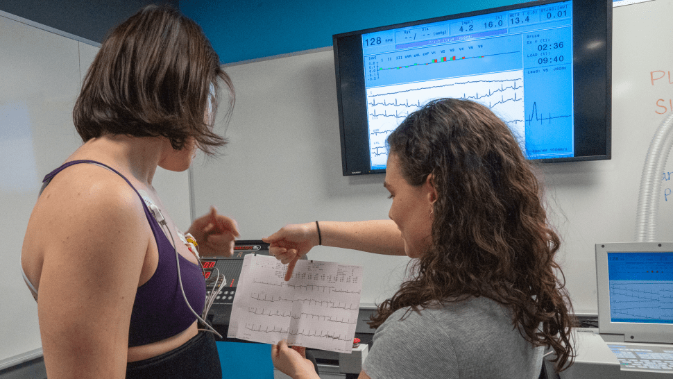 Two people look at a printout of data. One person is connected to a machine with wires and there is a screen with data on it.