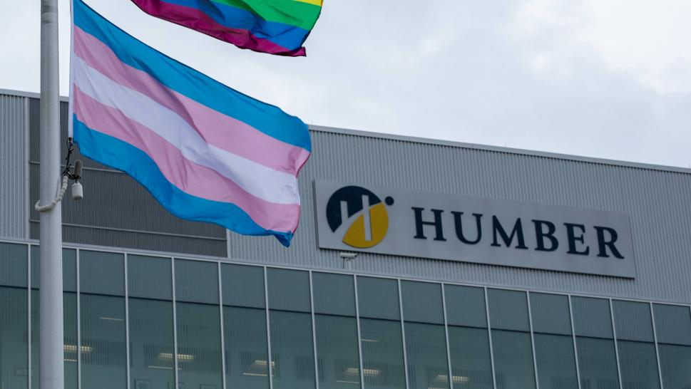 The Transgender Flag flies from a flag post. A building in the background has a sign that reads Humber.