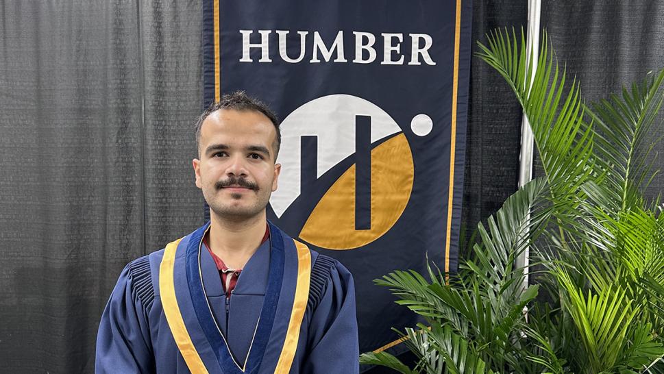 A person wearing convocation robes stands in front of a Humber Polytechnic banner.