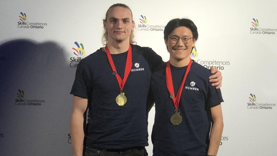 Two smiling Humber College students wear gold medals around their necks.