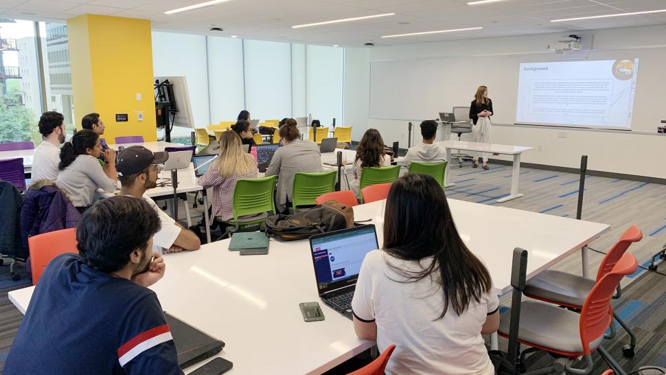 People sit in groups at tables while a person at the front of the room looks at a presentation on a screen.