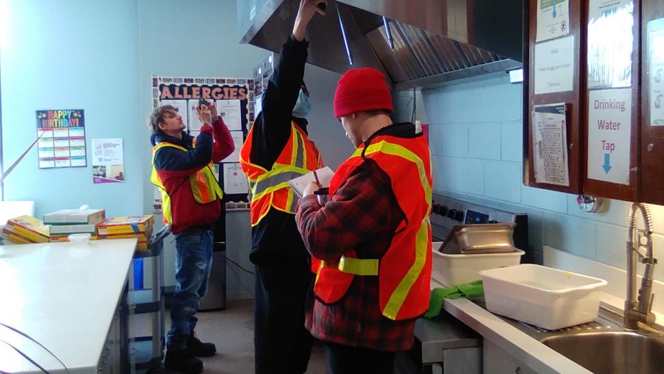 Students wearing safety vests take readings using a device inside a kitchen.