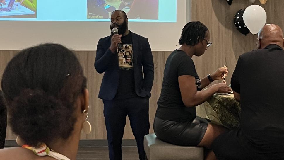 A person speaks into a microphone as others sitting at tables listen.