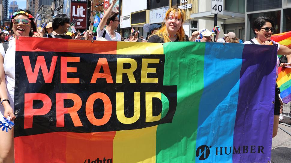 Students from Humber College participated in this year’s Toronto Pride Parade. People march in a parade while holding a rainbow-coloured banner reading We Are Proud and Humber.