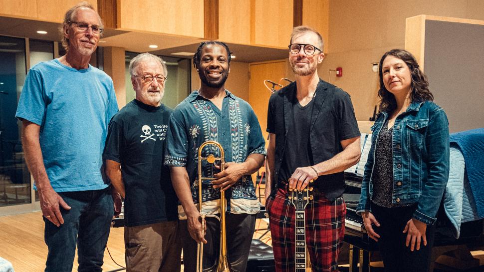 Five people stand in a row inside a recording studio. One person is holding a trombone and another is holding a guitar.