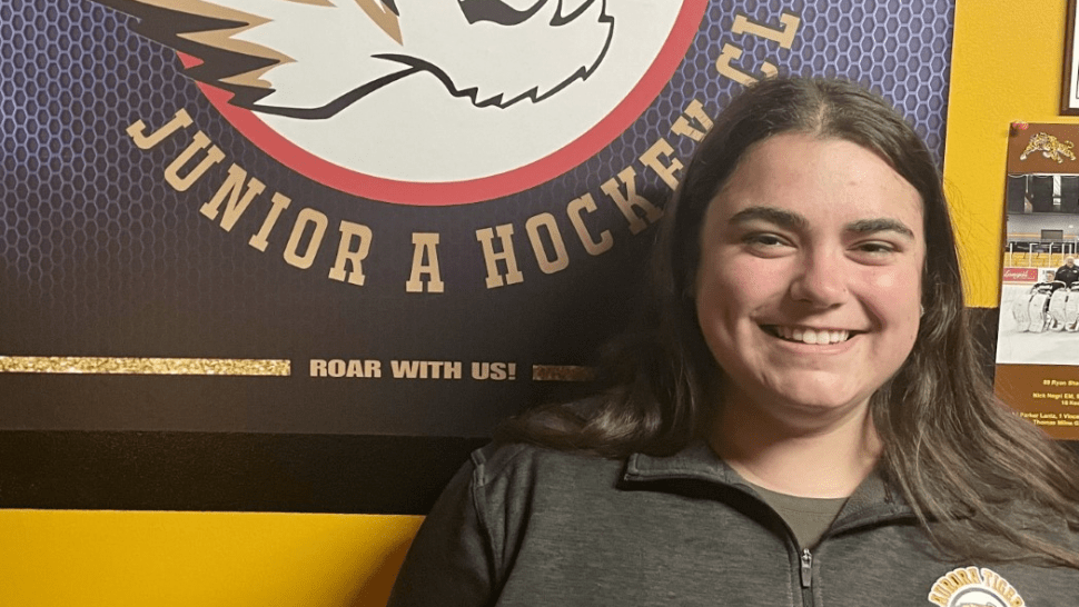 Humber Today is connecting with recent graduates such as Sierra Costa ahead of the Humber Alumni Weekend 2022. Sierra Costa stands beneath a sign that reads Aurora Tigers Junior A hockey club.