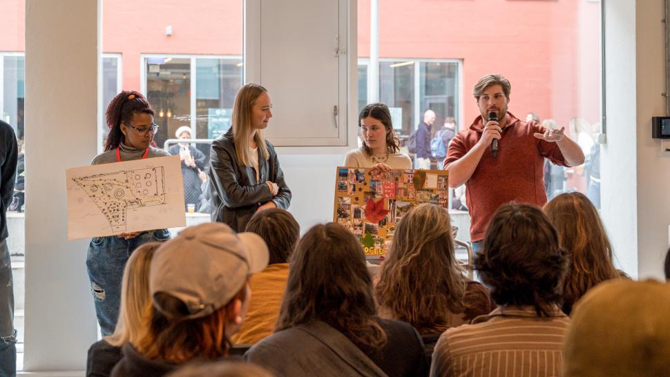 Several people give a presentation to a crowd at the KEA Charrette. One person is speaking into a microphone.