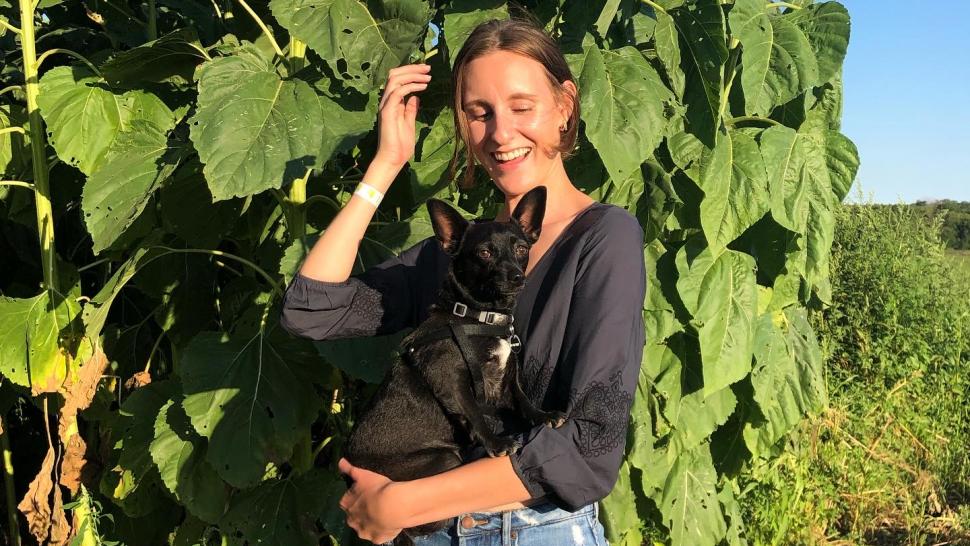 Erin Ross stands in front of tall green stalks, smiling and brushing her hair back with a small black dog in her arms