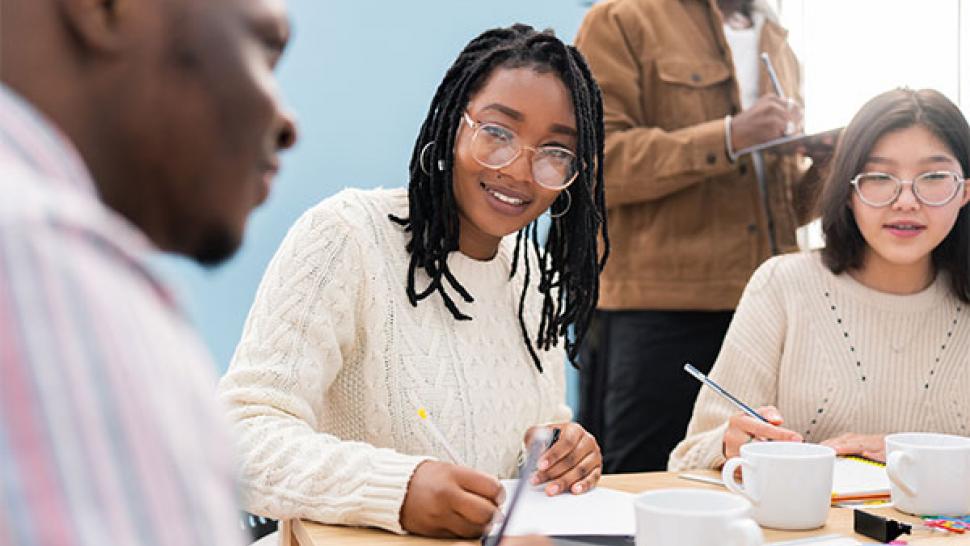 Three people sitting at a table are using pens to fill out applications. A fourth person stands in the background.