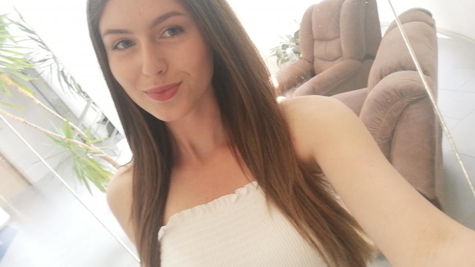 Aleksandra Peychinova smiles, closemouthed, wearing a white tank top in a selfie. She has long brown hair.