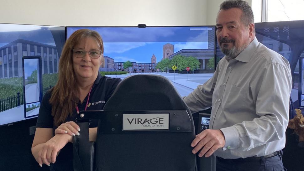 Humber’s Transportation Training Centre has a Virage truck simulator that’s being used as part of the Commercial Driver Training Class G to AZ Tractor Trailer course. Two people stand in front of a chair facing several screens with Virage Simulation on it.